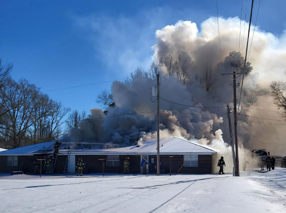 Large amount of smoke coming from apartment building with snow on the ground and firefighters fighting the fire.