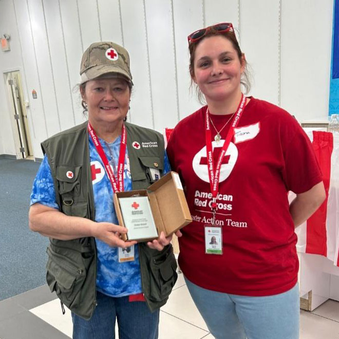 Cindye Schoof wearing a Red Cross hat, shirt and vest while holding a Red Cross award and standing next to a Red Cross employee.