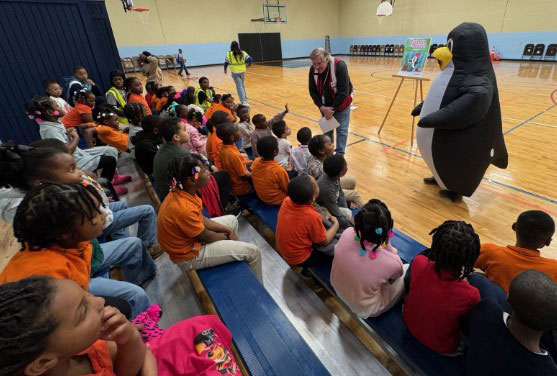 a Red Cross volunteer and Pedro the penguin mascot speak to a room full of school children.
