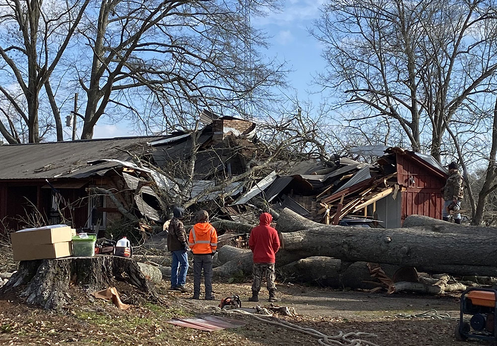 three people stading next to a damged house with a large oak tree that fell on top of it.