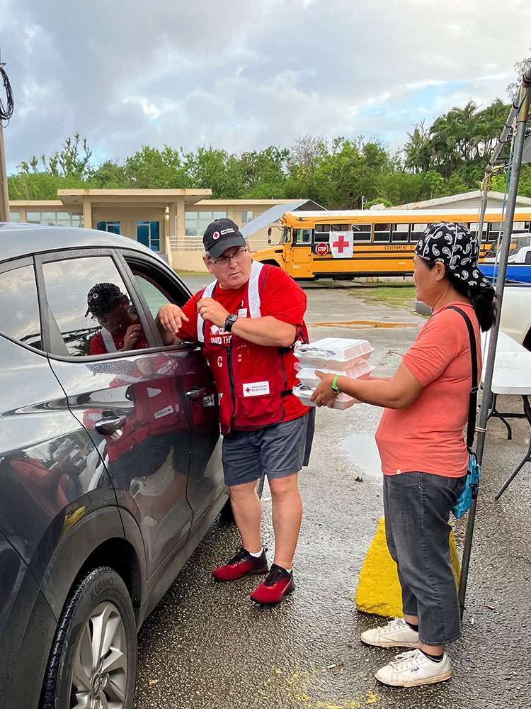 Ken Barger, in a Red Cross vest and hat, with a person next to him holding food in containers at a school parking lot.