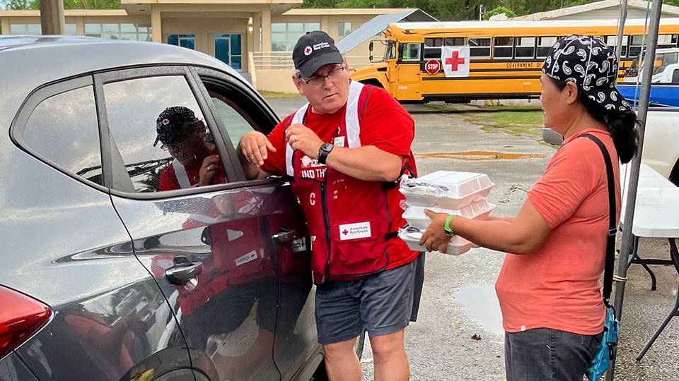 Ken Barger, in a Red Cross vestand hat, with a person next to him holding food in containers at a school parking lot.
