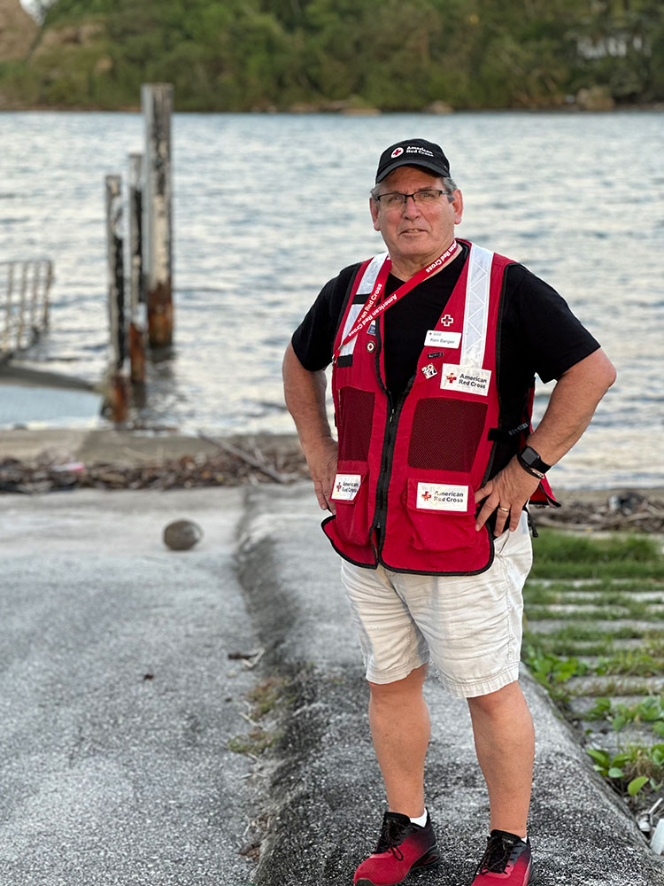 Ken Barger, in a Red Cross vest and hat, stands by a lake and smiles for picture.