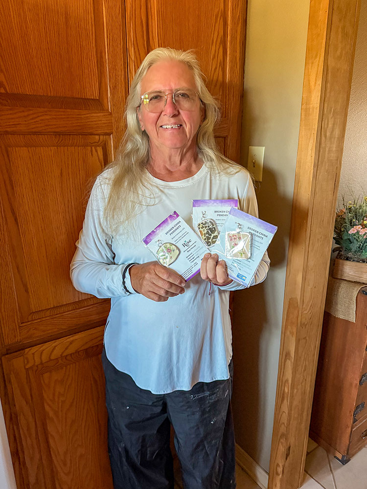 Kim Wilson wearing glasses and smiling while holding three packaged pendants.