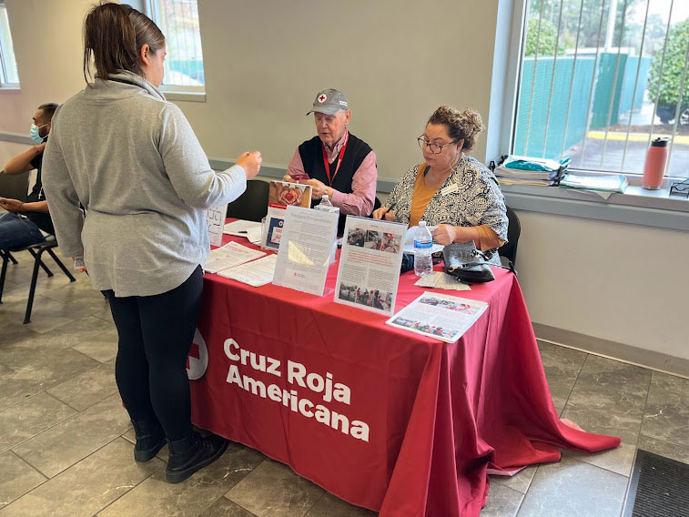 Red Cross volunteer standing in front of a table with Red Cross items and 2 Red Cross staff members.