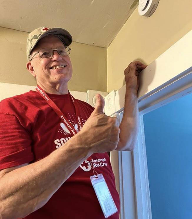 Norman Buescher wearing glasses, a Red Cross shirt and hat and giving a thumbs up.
