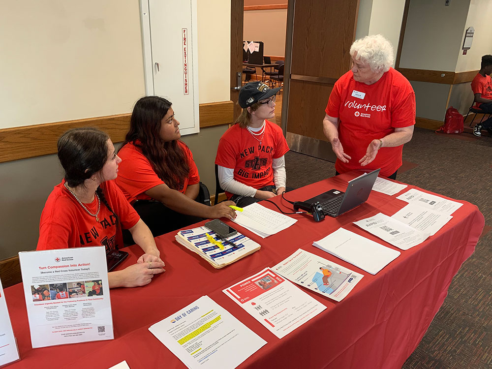 Patty Minga speaks to three young Red Cross volunteers that are sitting at a table with Red Cross pamphlets on it.