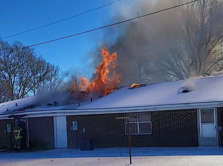 Flames coming from apartment building roof with snow on the ground and firefighters next to the building.