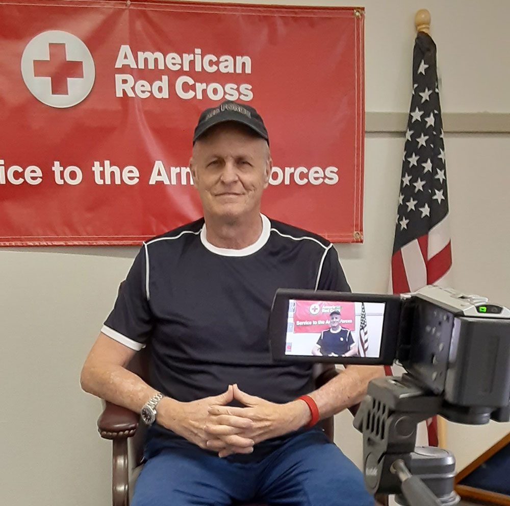 Eric Laurhammer in a blue shirt and cap sits in front of an American Red Cross banner, flanked by a U.S. flag. A video camera is recording him.