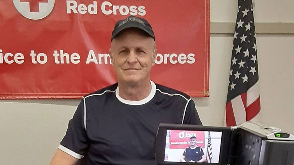 Eric Laurhammer in a blue shirt and cap sits in front of an American Red Cross banner, flanked by a U.S. flag. A video camera is recording him.