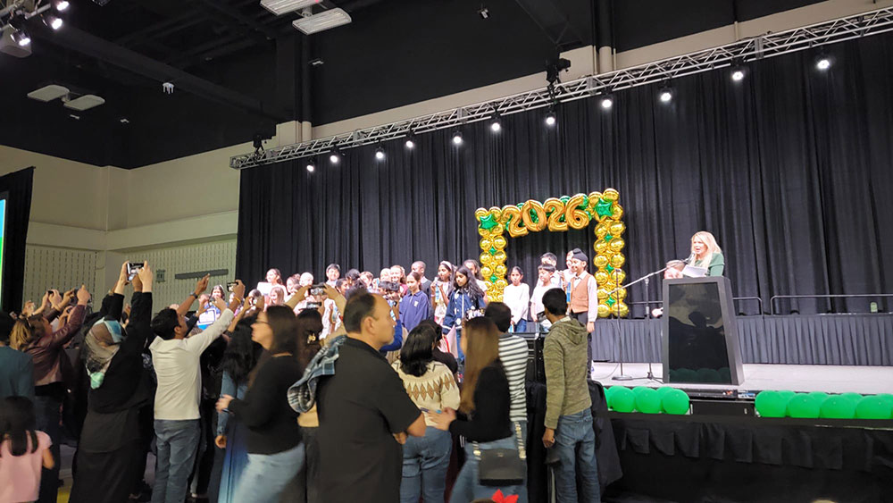 Group of people on stage with "2026" balloon backdrop, surrounded by audience taking photos