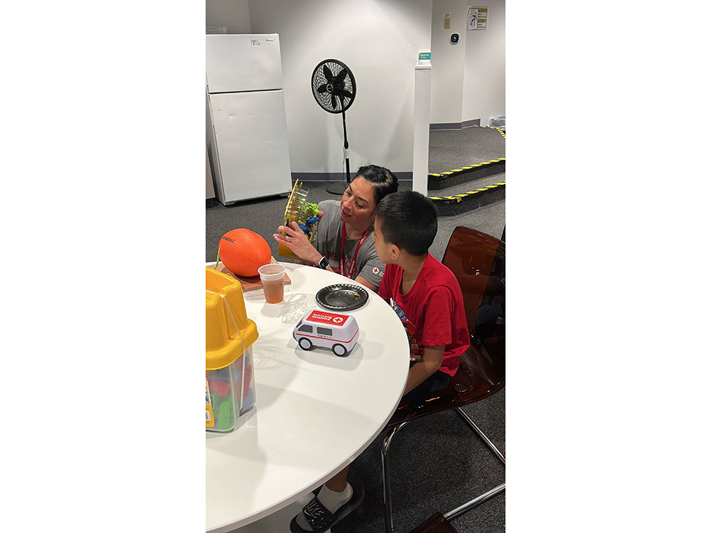 a Red Cross volunteer next to a child at a table and showing a boxed toy.