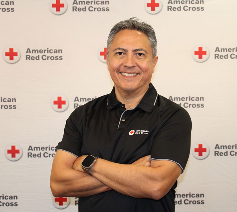 Javier Valdez, smiling with arms crossed, wearing a Red Cross shirt with a Red Cross backdrop behind him.