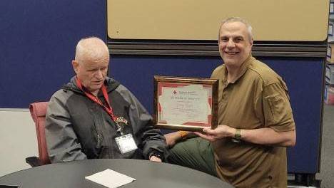 Red Cross volunteer Larry Roser sitting at a table with a cake and being presented a Red Cross service award.