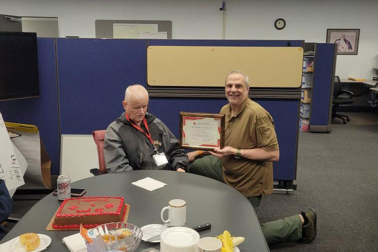 Red Cross volunteer Larry Roser sitting at a table with a cake and being presented a Red Cross service award.