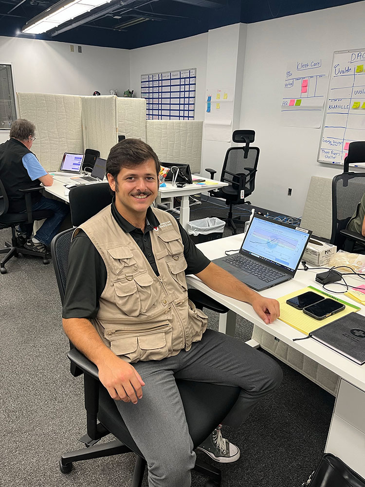 Nick Richardson wearing a Red Cross vest while sitting at a desk in an office with a laptop and smart phones.