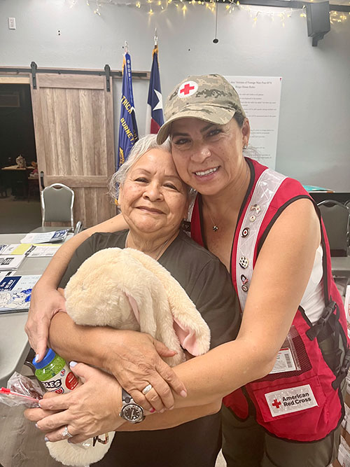 A Red Cross volunteer and an older woman embrace each other and smile for the camera.