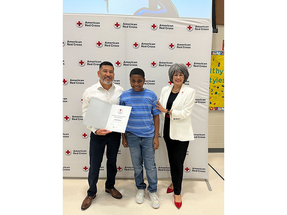 Three people stand smiling at an award event. The person on the left holds a certificate. They're in front of an American Red Cross backdrop.