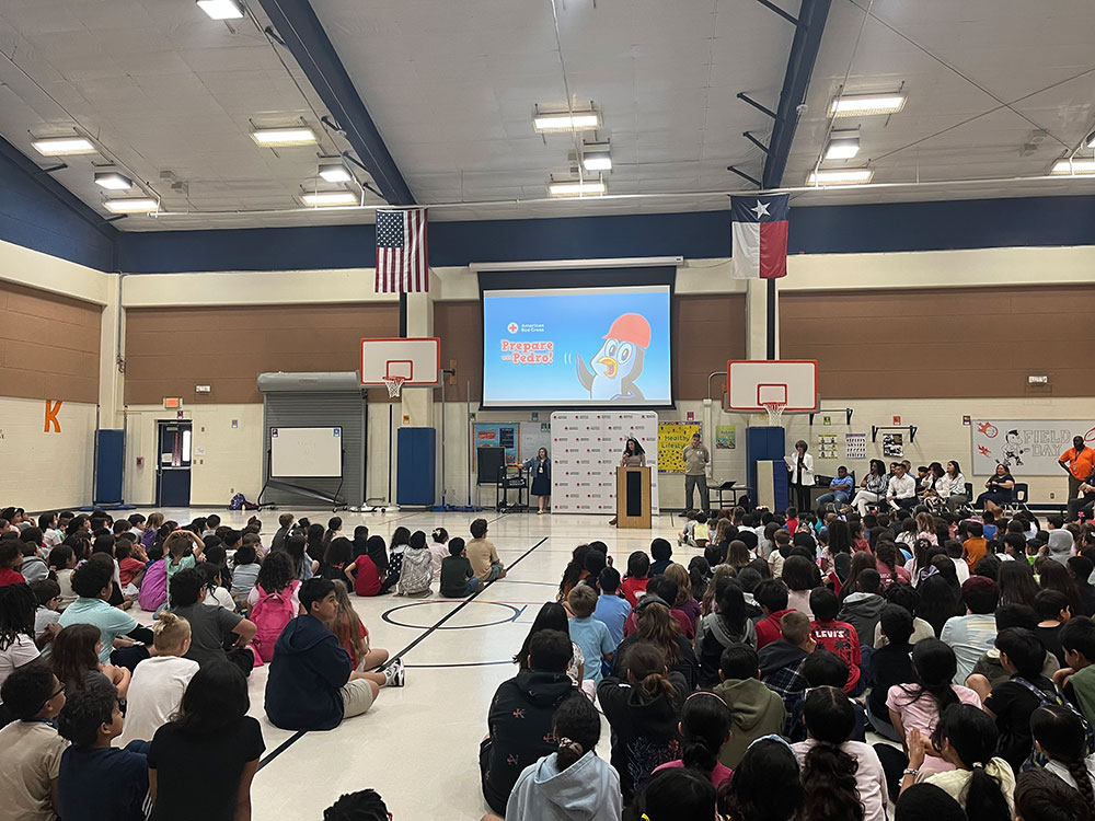 School assembly in a large gym with students seated on the floor. A person speaks at a podium; a screen displays an animation titled "Prepare with Pedro." American and Texan flags hang above.