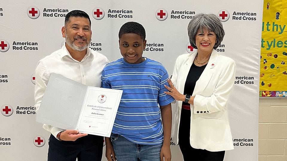 Three people stand smiling at an award event. The person on the left holds a certificate. They're in front of an American Red Cross backdrop.