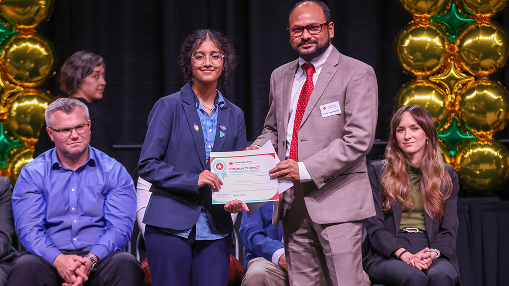 A smiling young person receives a certificate from a man in a suit at an event. They stand in front of gold balloons, with seated attendees in the background.