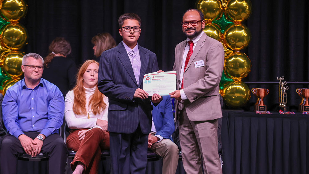 A young man in a suit receives an award certificate from an older man wearing a suit and red tie. They stand onstage, with others seated behind them.