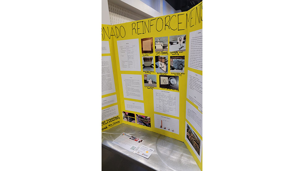 A yellow trifold science project display titled "Tornado Reinforcement" features text, charts, and photos. It is set up on a metallic table, conveying an educational tone.