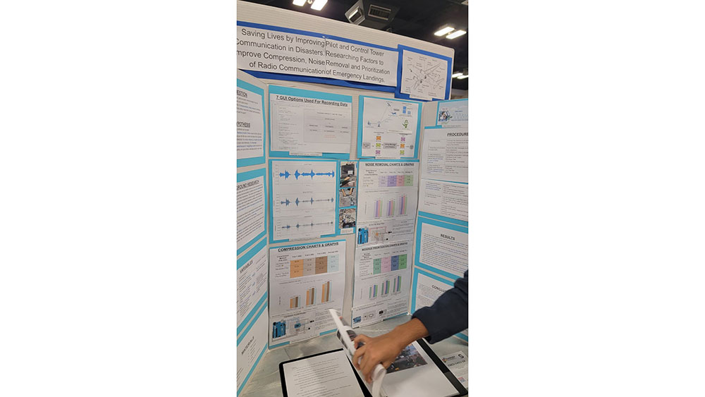A science fair project display board features graphs, charts, and text about improving pilot communication. A person is pointing to a page of notes.