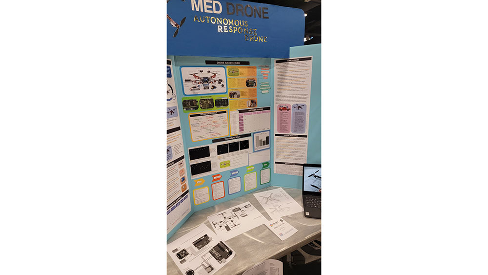 A science fair poster titled "Med Drone: Autonomous Response" displaying drone schematics, data charts, and information panels. Papers and a laptop are on the table.
