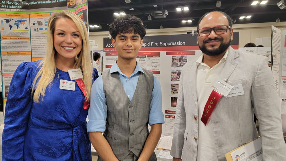 Three people at a science fair stand in front of project boards. They are smiling, wearing business attire with badges.