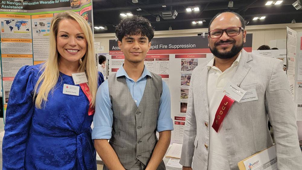 Three people at a science fair stand in front of project boards. They are smiling, wearing business attire with badges.