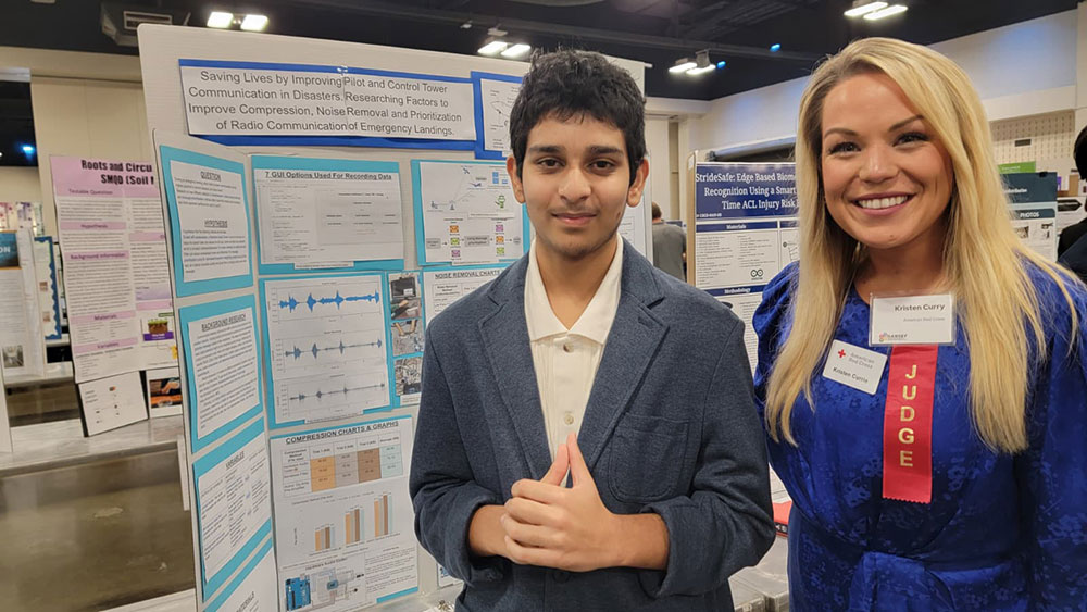 Two people at a science fair stand in front of project boards. They are smiling, wearing business attire with badges.