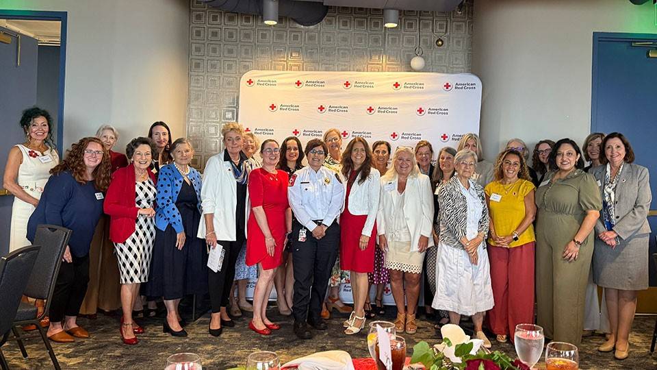 A group of women standing together in front of a Red Cross banner and smiling.