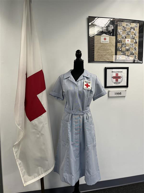 A vintage red cross nurse's dress on a mannequin beside a Red Cross flag.