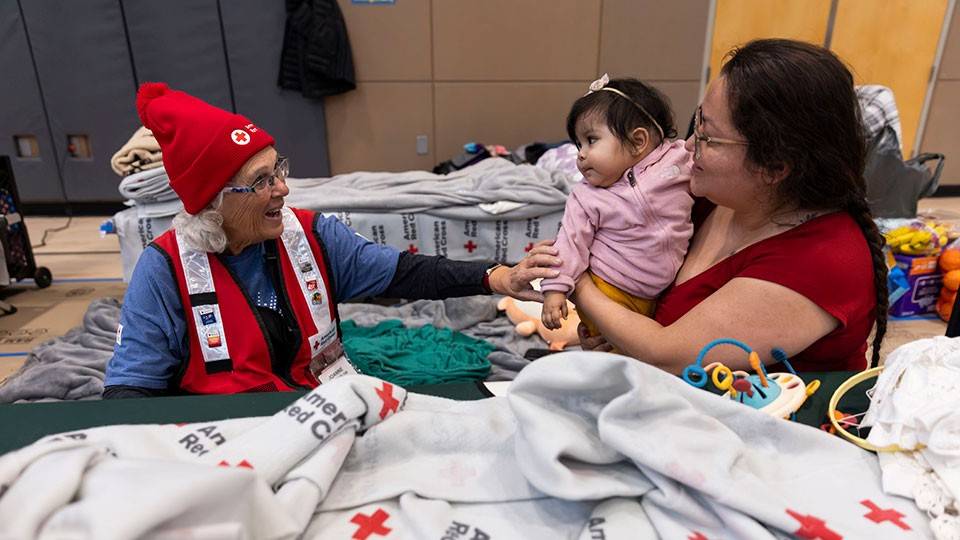 A Red Cross volunteer sitting with a mother and child in a Red Cross shelter.