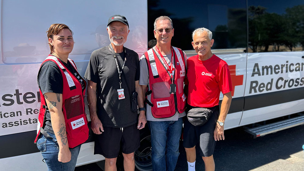 Four American Red Cross volunteers wearing red vests and badges on lanyards standing next to American Red Cross vehicle .