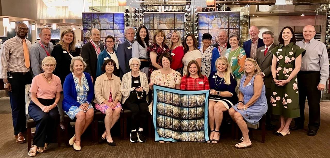 a group of people both sitting and standing posing with a quilt