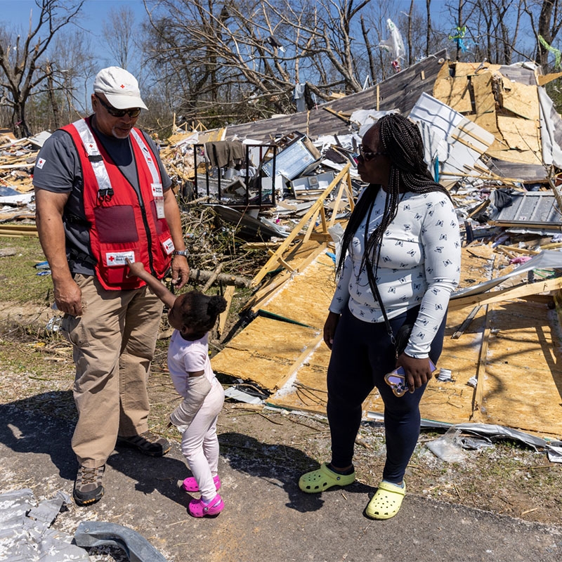 Red Cross volunteer with womand and child next to damaged buildings.