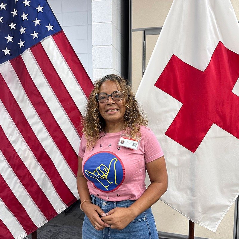 Red Cross volunteer standing in front of American and Red Cross flags smiling for camera.