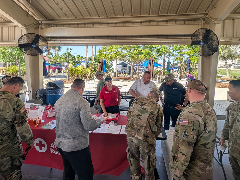 military members in camo at a Red Cross table picking up paperwork and other Red Cross items.