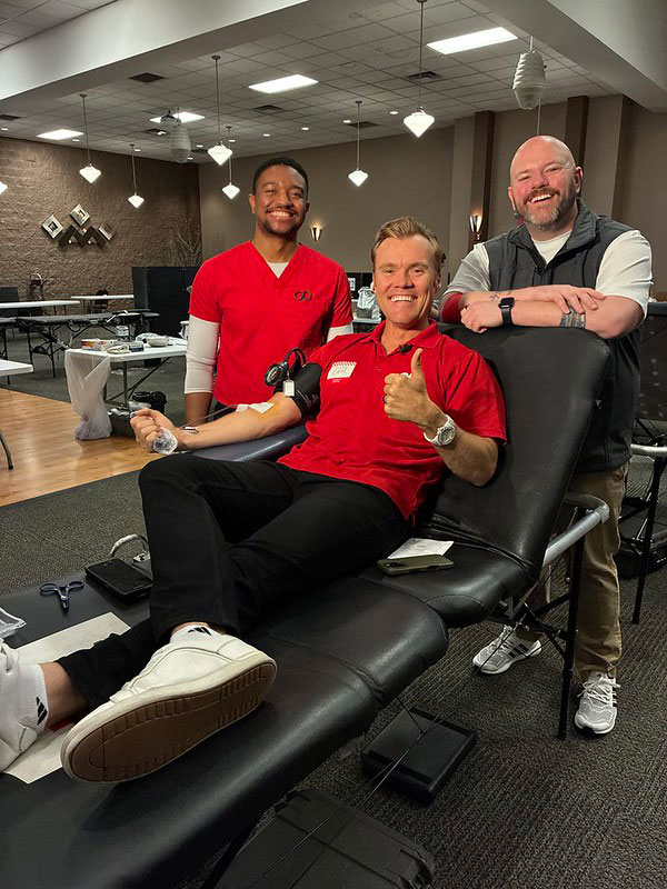 a person sitting in a chair donating blood while smiling and giving a thumbs up. Two other people stand next to him.