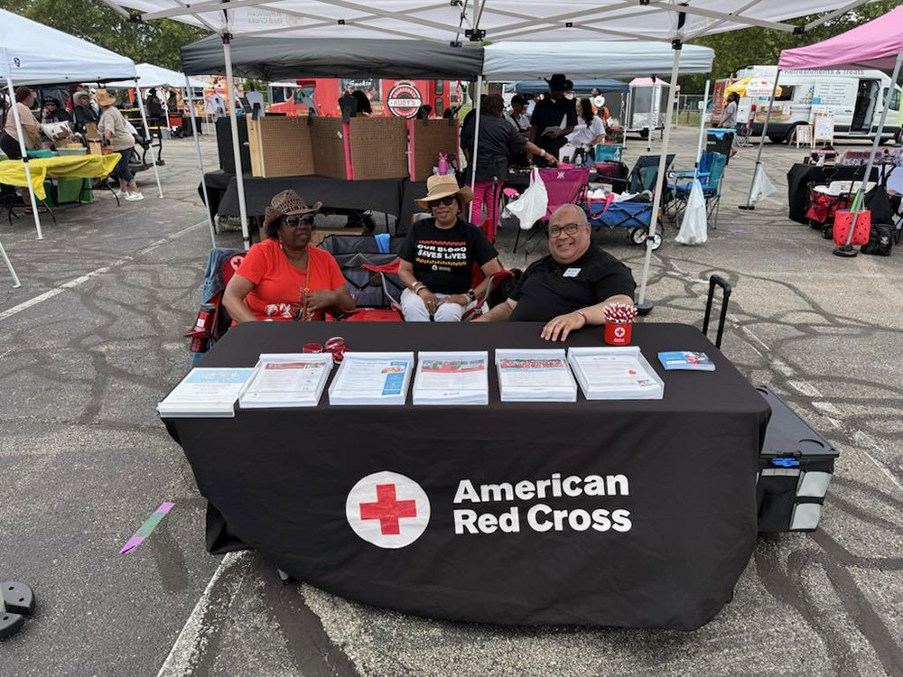a table with a Red Cross table cloth and Red Cross promotional items in a parking lot with three people behind the table..