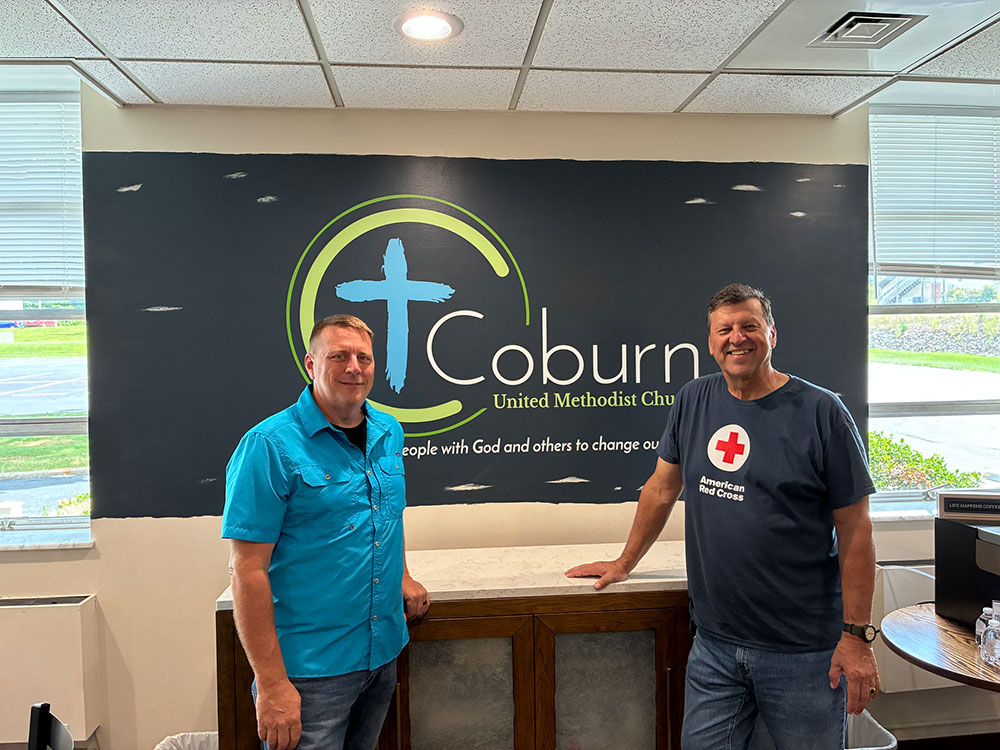 two people smiling and standing in front of a Coburn United Methodist Church sign inside a room.
