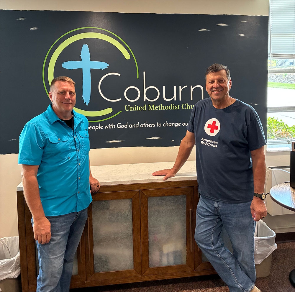 two people smiling and standing in front of a Coburn United Methodist Church sign inside a room.