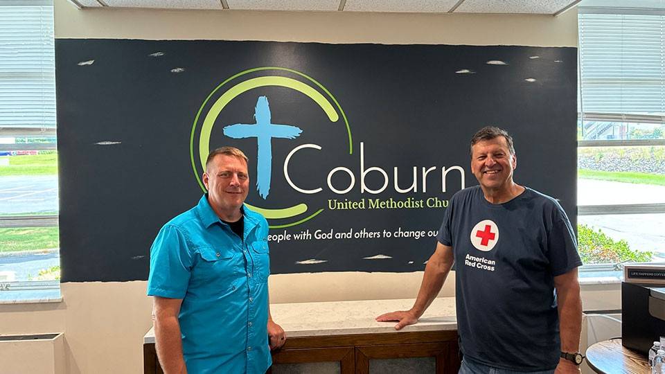 two people smiling and standing in front of a Coburn United Methodist Church sign inside a room.