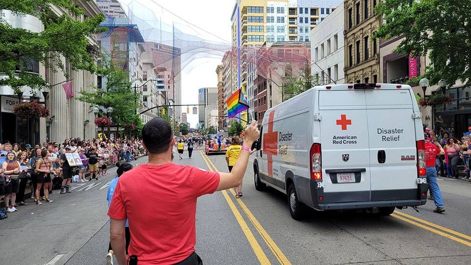 a pride parade with a large crowd lining a city street and a red cross van.