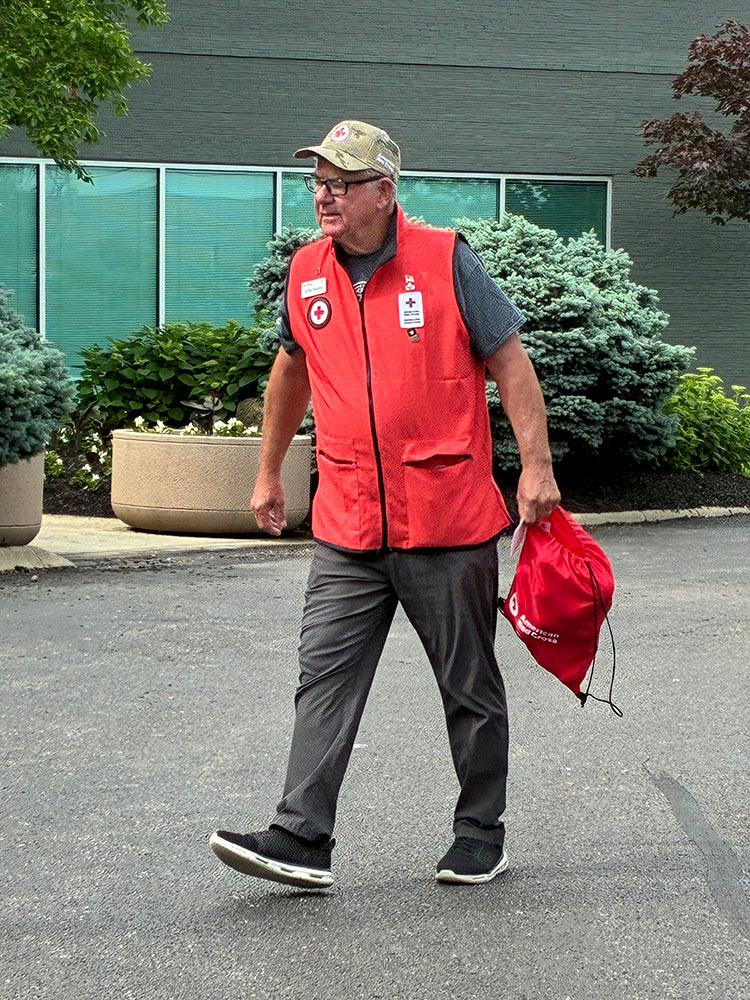 Jeff Earley wearing a Red Cross vest and holding a Red Cross bag while walking on the pavement.