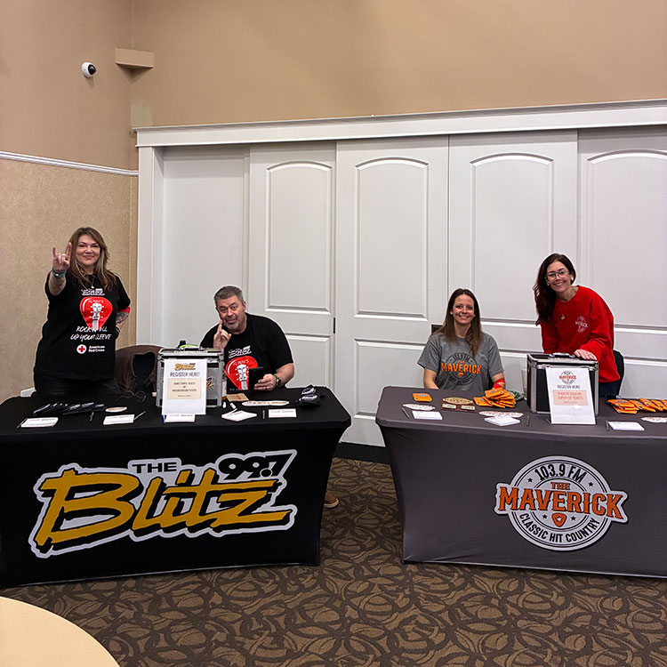 four people behind tables in a room that have the Blitz 99.7 and The Maverick 103.9 logos. Promotional items are on the tables.