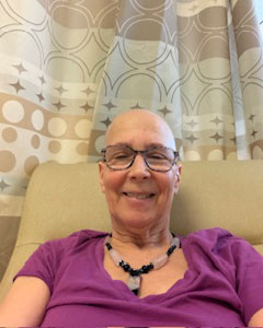 Norma Wasserstrom sitting in chair wearing glasses a necklace and smiling.
