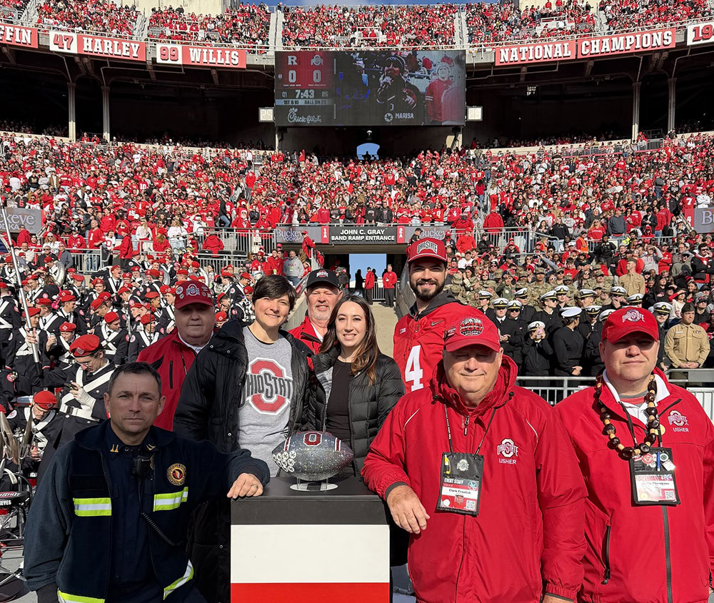 Ohio state football staff, paramedic, and Red Cross volunteers stand next to the Ohio state football trophy in the Ohio state football stadium that's filled with fans.
